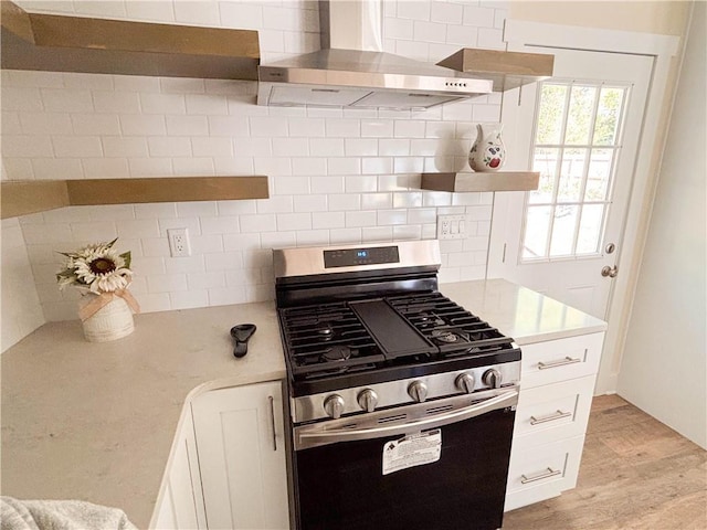 kitchen featuring tasteful backsplash, white cabinetry, stainless steel range with gas stovetop, wall chimney range hood, and light wood-type flooring