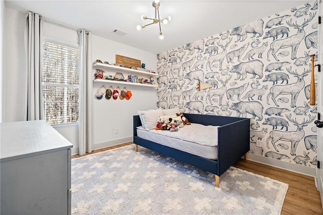 bedroom featuring wood finished floors, wallpapered walls, baseboards, a chandelier, and an accent wall