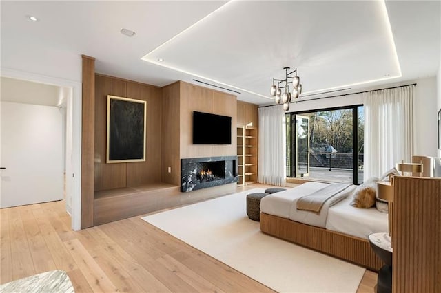 bedroom featuring light wood-type flooring, a raised ceiling, a warm lit fireplace, and a chandelier