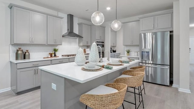 kitchen with a center island with sink, appliances with stainless steel finishes, gray cabinetry, a kitchen breakfast bar, and wall chimney range hood