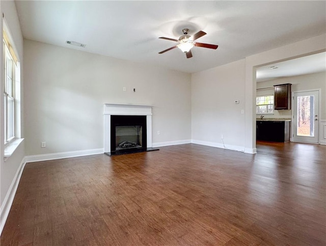 unfurnished living room with visible vents, a glass covered fireplace, dark wood finished floors, baseboards, and ceiling fan