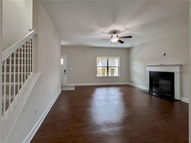 unfurnished living room with stairway, baseboards, dark wood finished floors, ceiling fan, and a glass covered fireplace