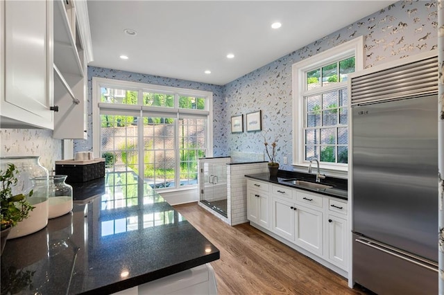 kitchen with stainless steel built in refrigerator, a sink, wallpapered walls, white cabinetry, and wood finished floors