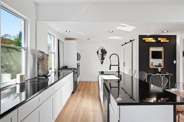 kitchen featuring light wood finished floors, black gas stove, a center island with sink, a barn door, and white cabinetry