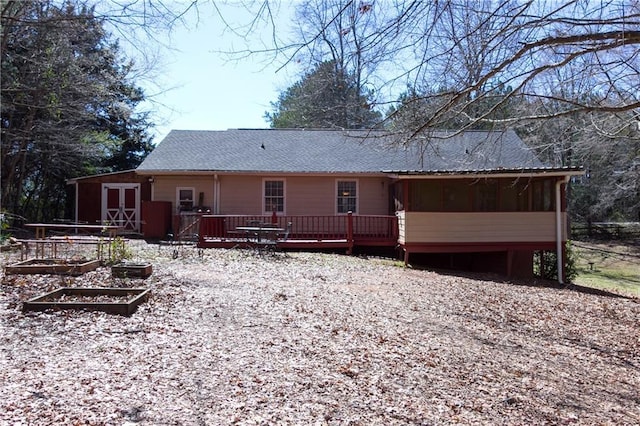 rear view of property featuring a wooden deck, a vegetable garden, a sunroom, roof with shingles, and an outdoor structure