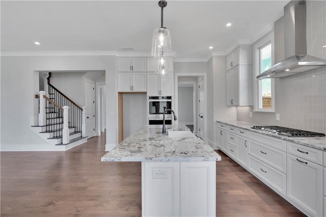 kitchen with white cabinets, appliances with stainless steel finishes, wall chimney exhaust hood, an island with sink, and hanging light fixtures