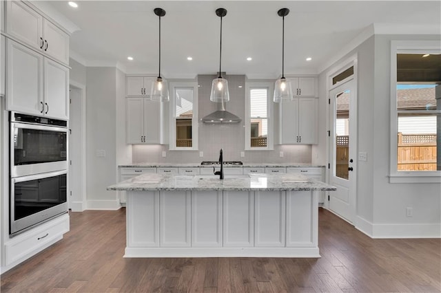 kitchen with stainless steel double oven, a center island with sink, decorative backsplash, white cabinets, and light stone counters