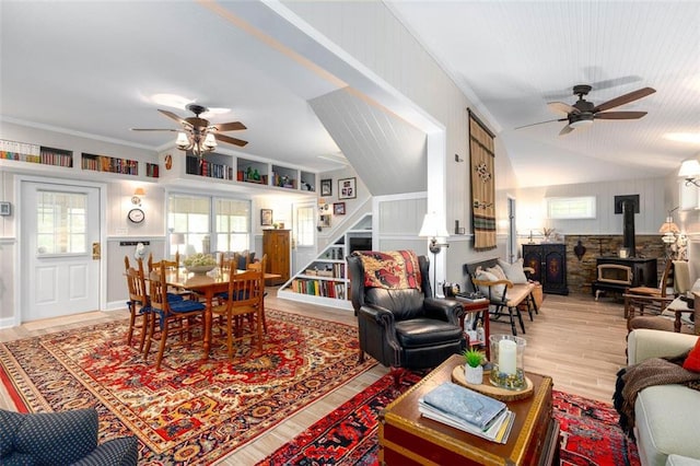 living room with ceiling fan, a wood stove, light hardwood / wood-style flooring, crown molding, and vaulted ceiling