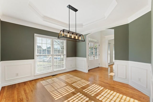 unfurnished dining area with a wainscoted wall, ornamental molding, a raised ceiling, and light wood-style floors