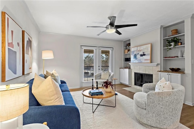 living room with built in shelves, ceiling fan, a tiled fireplace, and hardwood / wood-style flooring