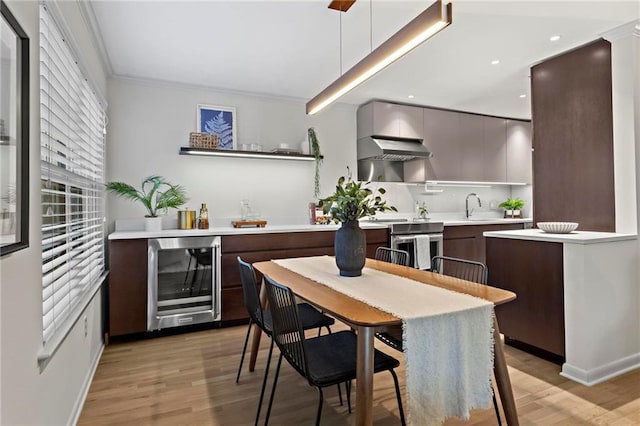 kitchen with dark brown cabinetry, beverage cooler, crown molding, wall chimney range hood, and hanging light fixtures