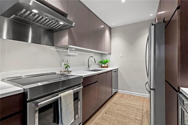 kitchen with dark brown cabinets, stainless steel appliances, wall chimney range hood, sink, and light hardwood / wood-style flooring