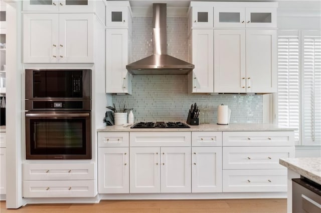 kitchen with wall chimney range hood, tasteful backsplash, light stone counters, oven, and white cabinets