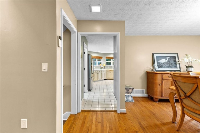 hall with light wood-type flooring, visible vents, a textured ceiling, and baseboards