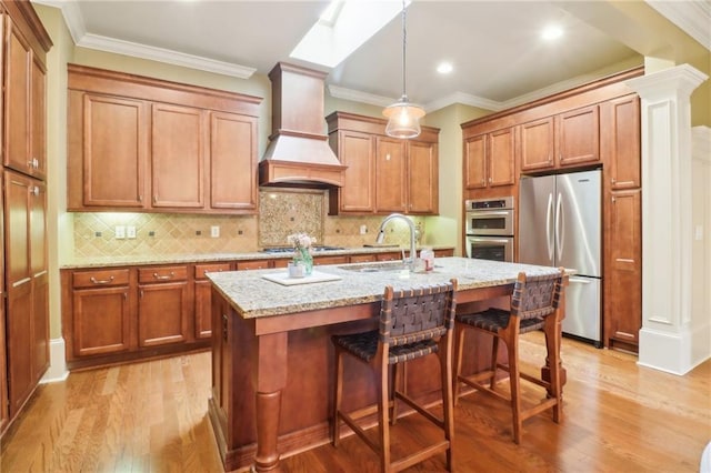 kitchen with sink, custom exhaust hood, light stone counters, stainless steel appliances, and a center island with sink