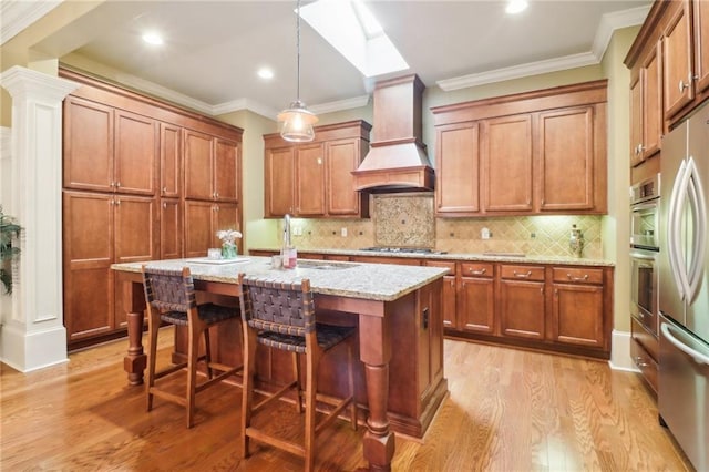 kitchen with custom exhaust hood, appliances with stainless steel finishes, pendant lighting, light stone countertops, and a kitchen island with sink