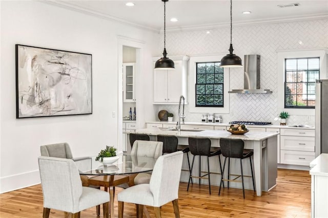 kitchen with a center island with sink, ornamental molding, white cabinetry, a sink, and wall chimney exhaust hood