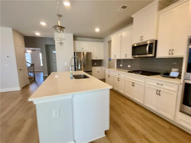 kitchen with light hardwood / wood-style flooring, appliances with stainless steel finishes, a kitchen island with sink, white cabinets, and decorative light fixtures