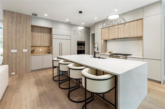 kitchen featuring an island with sink, light brown cabinetry, light wood-type flooring, sink, and decorative light fixtures