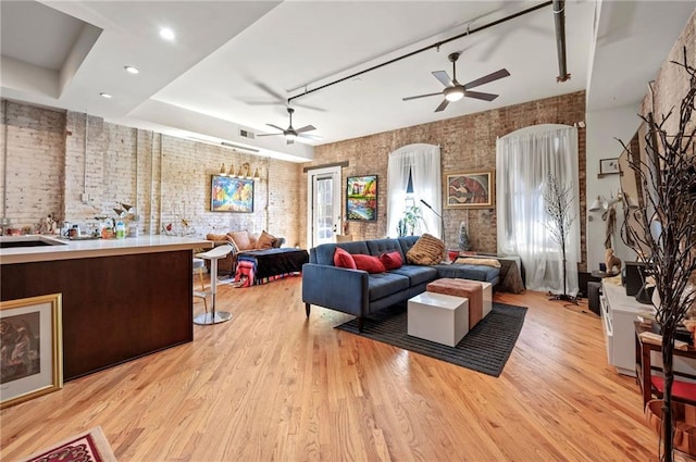 living room with ceiling fan, light wood-type flooring, a raised ceiling, and brick wall