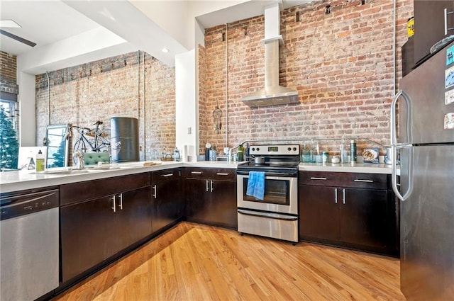 kitchen featuring brick wall, light wood finished floors, stainless steel appliances, light countertops, and wall chimney range hood