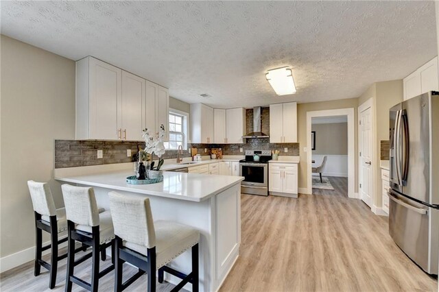 kitchen with white cabinetry, a kitchen breakfast bar, kitchen peninsula, stainless steel appliances, and wall chimney range hood