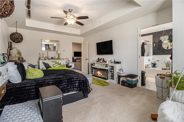 carpeted bedroom with a ceiling fan, a tray ceiling, a lit fireplace, and ensuite bathroom