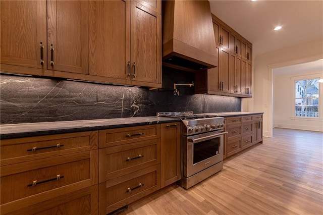 kitchen with brown cabinets, custom exhaust hood, light wood-style flooring, backsplash, and high end range