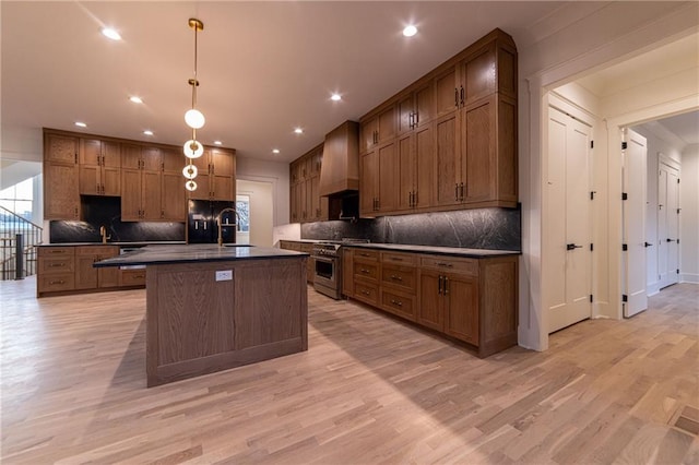 kitchen with stainless steel range, dark countertops, custom range hood, a kitchen island with sink, and a sink