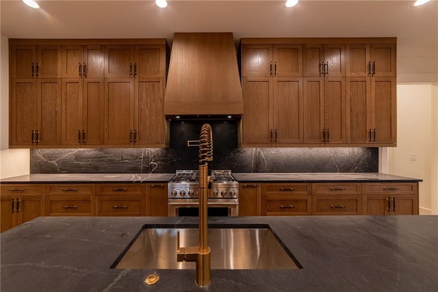 kitchen with dark stone counters, stainless steel range, custom exhaust hood, and brown cabinetry