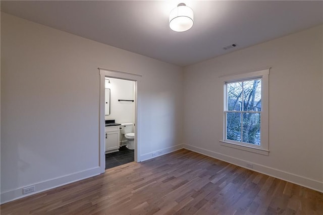 unfurnished bedroom featuring dark wood-style flooring, ensuite bath, visible vents, and baseboards