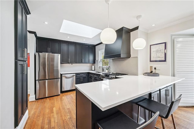 kitchen with a sink, a peninsula, a skylight, stainless steel appliances, and wall chimney exhaust hood