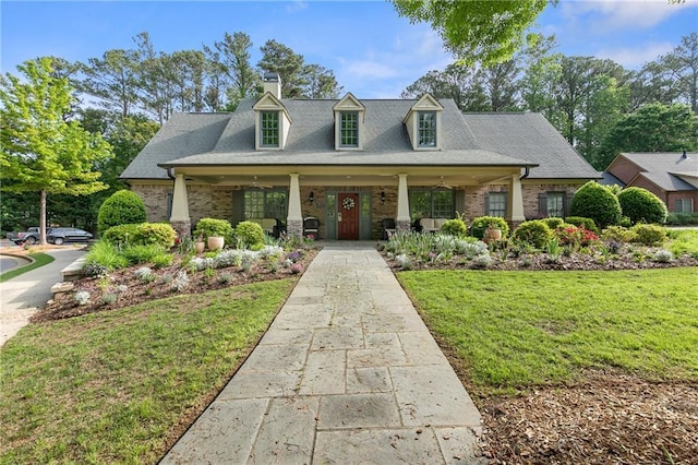 cape cod home featuring covered porch, a chimney, a front yard, roof with shingles, and brick siding