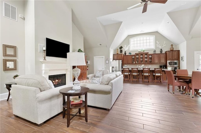 living area with visible vents, a glass covered fireplace, light wood-style floors, high vaulted ceiling, and a ceiling fan