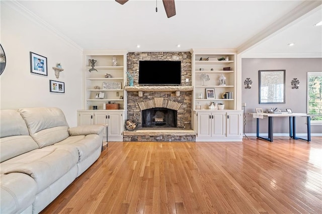 living area with built in shelves, light wood-style flooring, ceiling fan, a stone fireplace, and ornamental molding