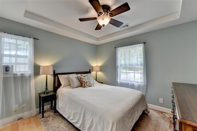 bedroom featuring a raised ceiling, a ceiling fan, baseboards, wood finished floors, and visible vents