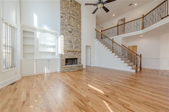 unfurnished living room featuring a stone fireplace, a towering ceiling, ceiling fan, light hardwood / wood-style floors, and built in shelves