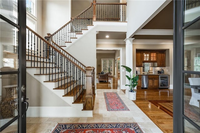 foyer with beverage cooler, a towering ceiling, baseboards, decorative columns, and crown molding