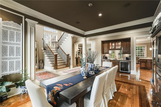 dining room featuring beverage cooler, light wood-style flooring, stairway, ornate columns, and indoor wet bar