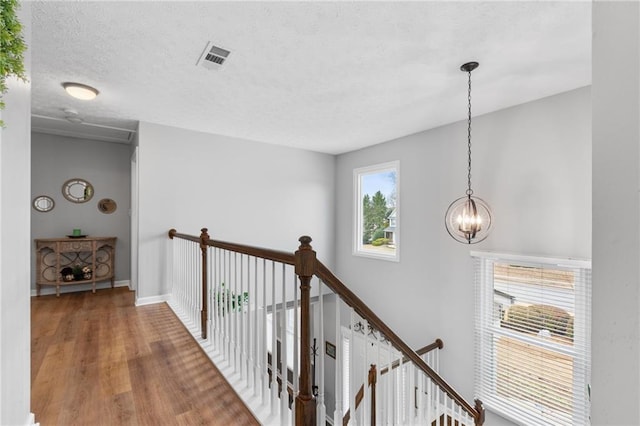 hallway with a textured ceiling, wood finished floors, visible vents, an upstairs landing, and an inviting chandelier