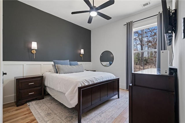 bedroom featuring visible vents, ceiling fan, light wood-style floors, and a wainscoted wall