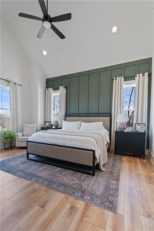 bedroom with a decorative wall, multiple windows, light wood-style floors, and high vaulted ceiling