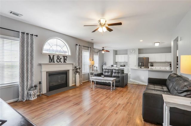 living area featuring a ceiling fan, light wood finished floors, a glass covered fireplace, and visible vents
