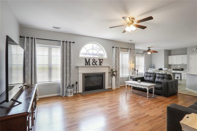 living area featuring baseboards, light wood finished floors, a glass covered fireplace, ceiling fan, and visible vents