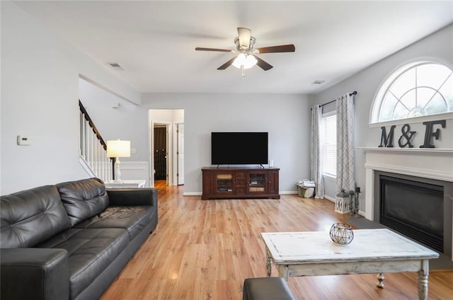 living area with light wood finished floors, a glass covered fireplace, ceiling fan, visible vents, and stairs