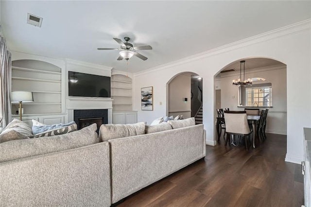 living room with built in features, ceiling fan with notable chandelier, crown molding, and dark wood-type flooring