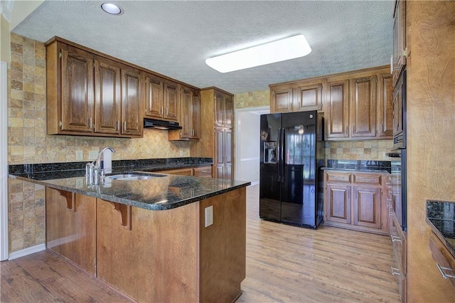 kitchen with light wood-style floors, black fridge with ice dispenser, a peninsula, under cabinet range hood, and a sink