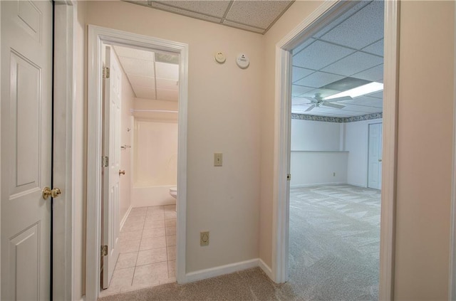 hallway featuring baseboards, a paneled ceiling, and light colored carpet