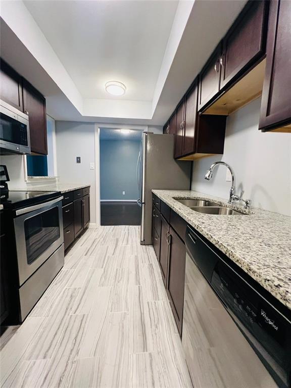kitchen with a tray ceiling, dark brown cabinetry, a sink, light stone countertops, and appliances with stainless steel finishes