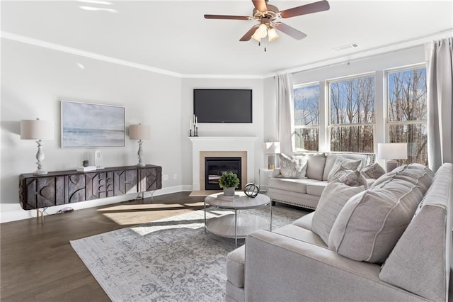 living area with a ceiling fan, baseboards, ornamental molding, dark wood-style floors, and a glass covered fireplace